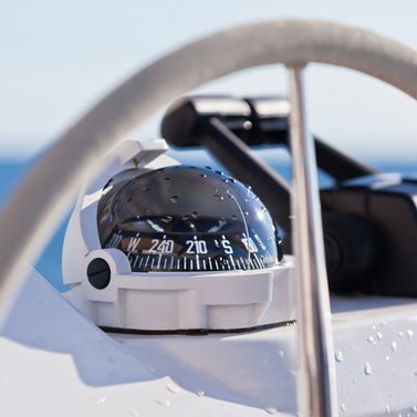 Zeigt einen Kompass, Teile eines Steuerrads sowie den Gashebel eines Bootes. 